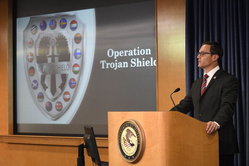 Randy Grossman speaks at a podium with a Justice Department logo, near a screen with the words "Operation Trojan Shield."