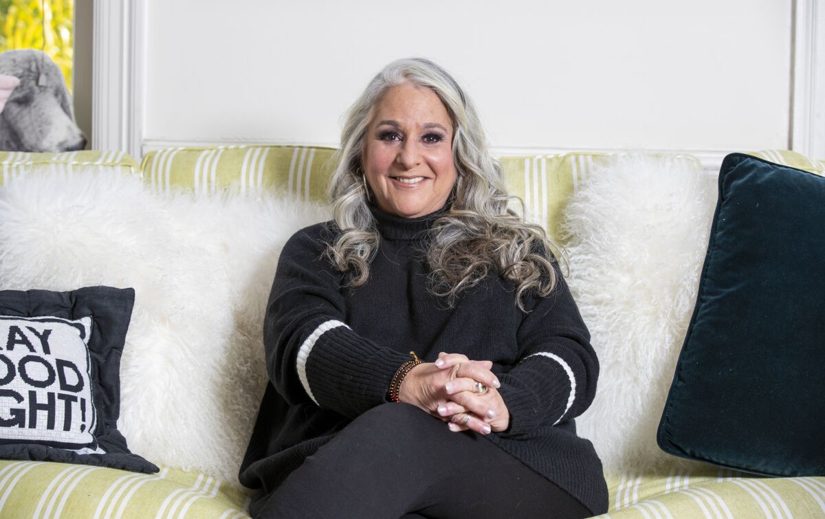 A woman with long gray hair, wearing black, sitting on a couch