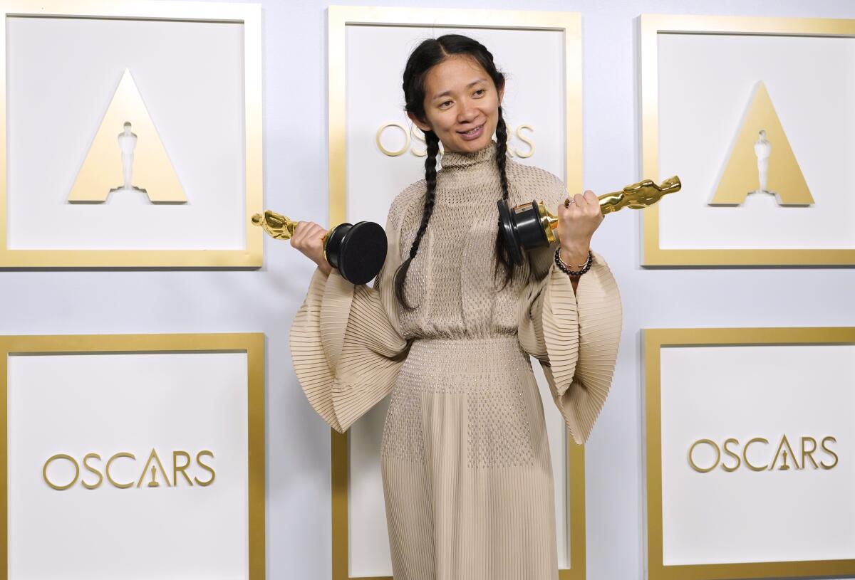 Chloé Zhao, in a gown with flowing sleeves, smiles as she holds an Oscar statuette in each hand.