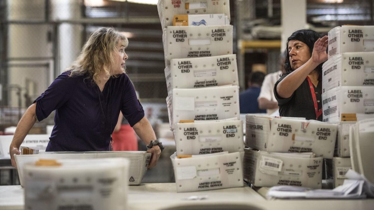 Orange County Registrar of Voters employees Barbara Strauss, left, and Hilda Rodriguez grab trays of mail-in ballots to sort through Wednesday at their facility in Santa Ana.