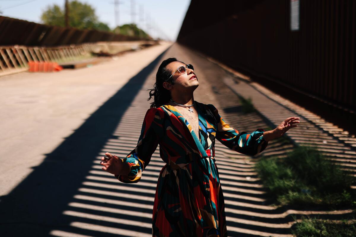 A transgender person in sunglasses and a colorful dress poses in front of a border wall with Mexico. 