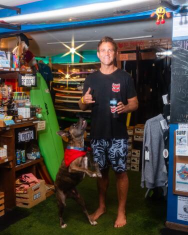 A dog stands next to a man in a black T-shirt and blue board shorts, who gives a hang-loose hand gesture, in a surf shop
