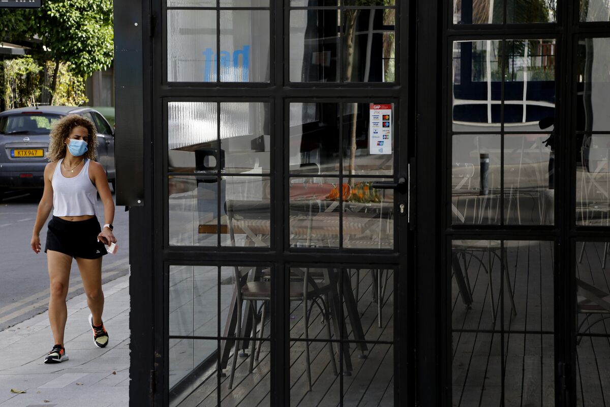 A woman wearing protective face mask walks outside of a closed cafe amid COVID-19 pandemic restrictions government in central capital Nicosia, Cyprus, Thursday, May 6, 2021. Cyprus has unveiled a phased rollback of COVID-19 lockdown restrictions over the next month including a shortened curfew and a reopening of all schools next week, but will enforce the compulsory display of proof of vaccination, virus testing or convalescence from the disease in areas were people gather in numbers, including restaurants and churches. (AP Photo/Petros Karadjias)