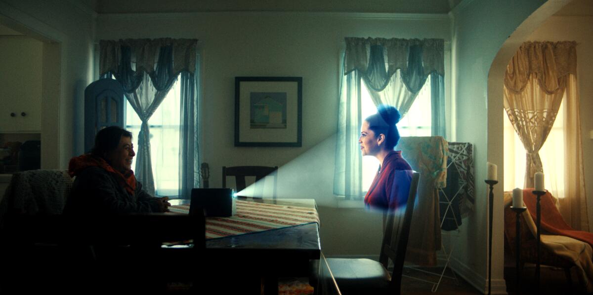 A woman sits at a table talking to a hologram of her daughter.