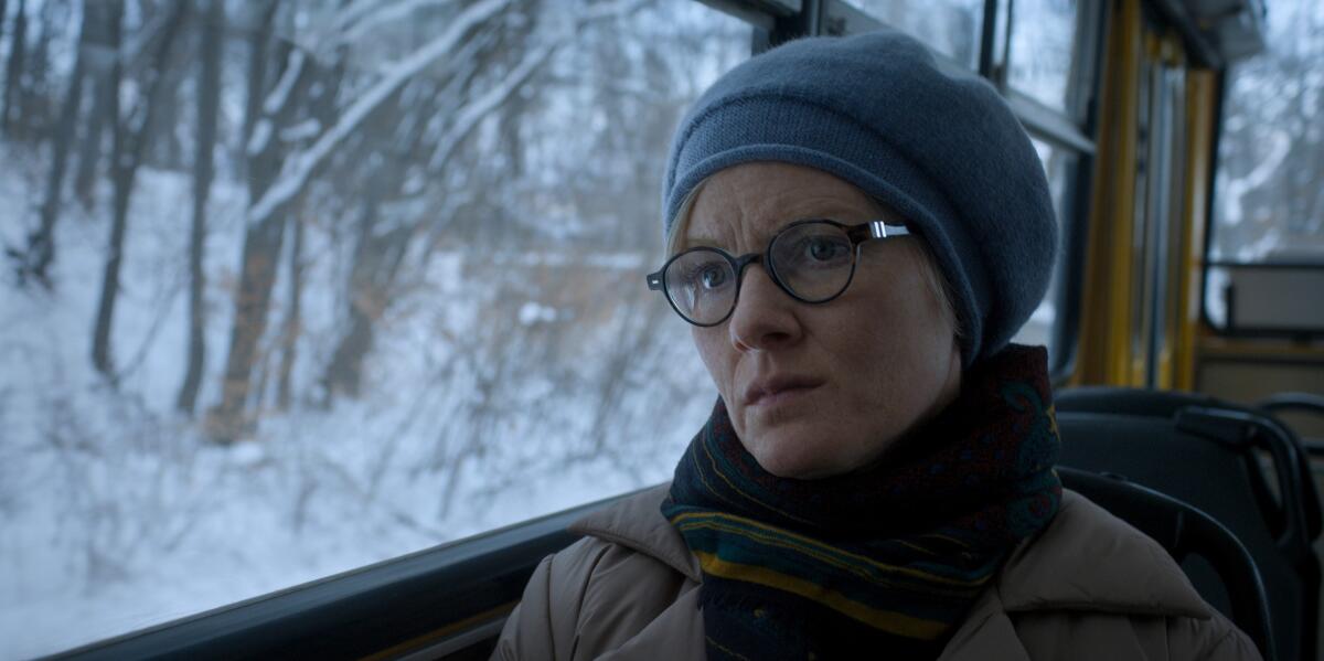 A woman in a gray hat, scarf and glasses sits in a car that's moving past a snowy forest.