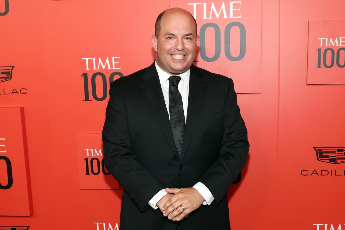 A bald man wearing a black suit and tie stands in front of a red wall emblazoned with "Time 100."