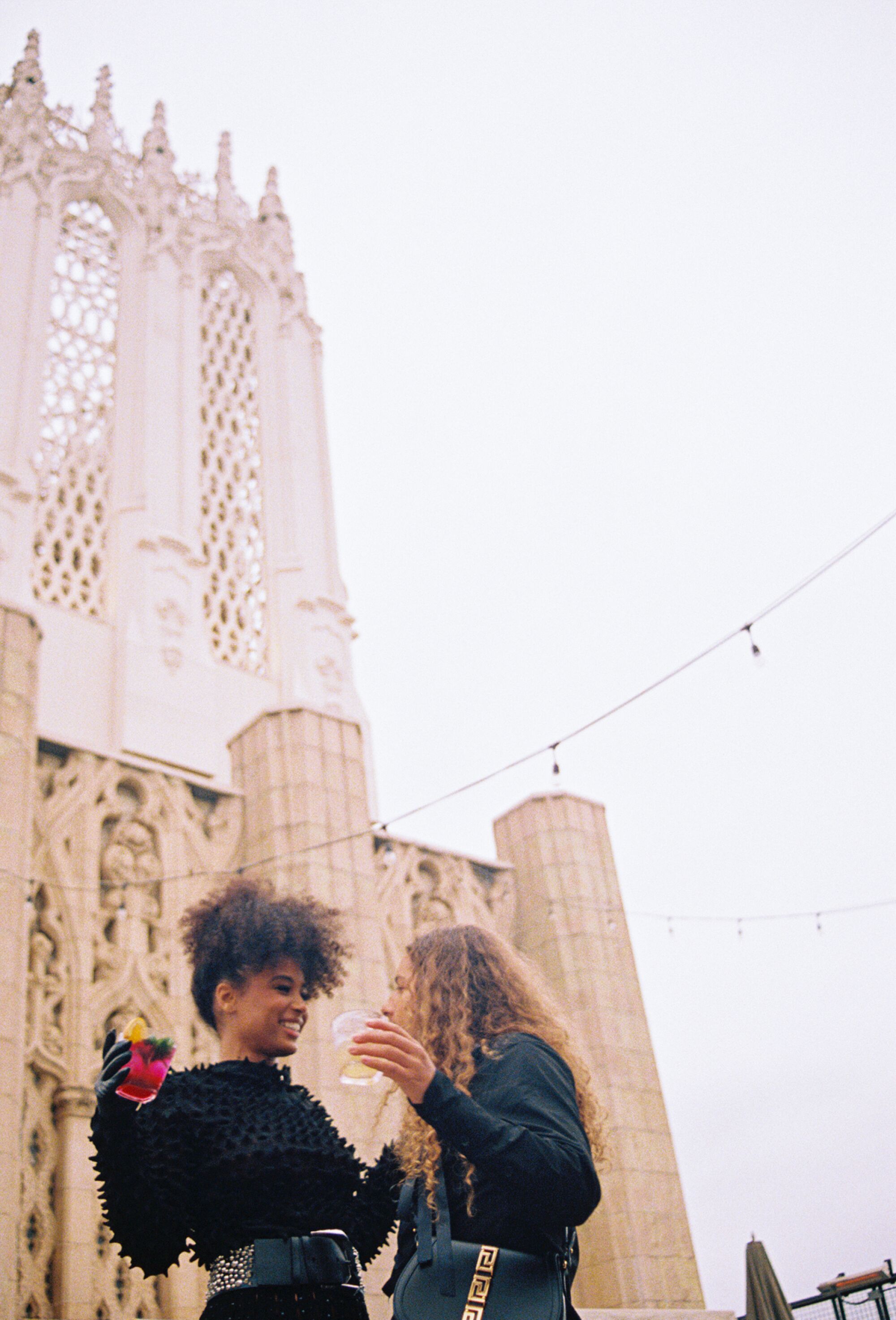 Two people wearing black laugh and hold their drinks on a rooftop. 