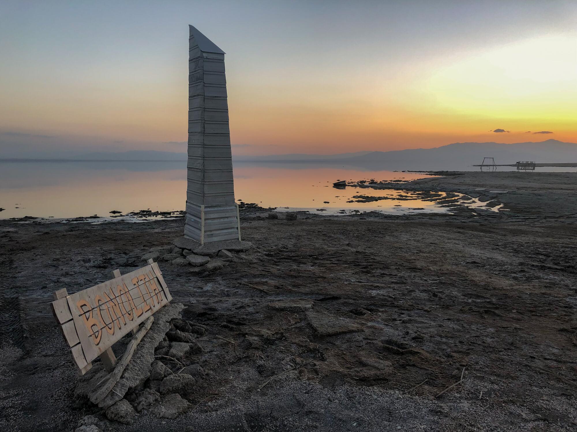 A sunset scene on a beach with a sign reading "Bonolith" and a wooden monolith figure