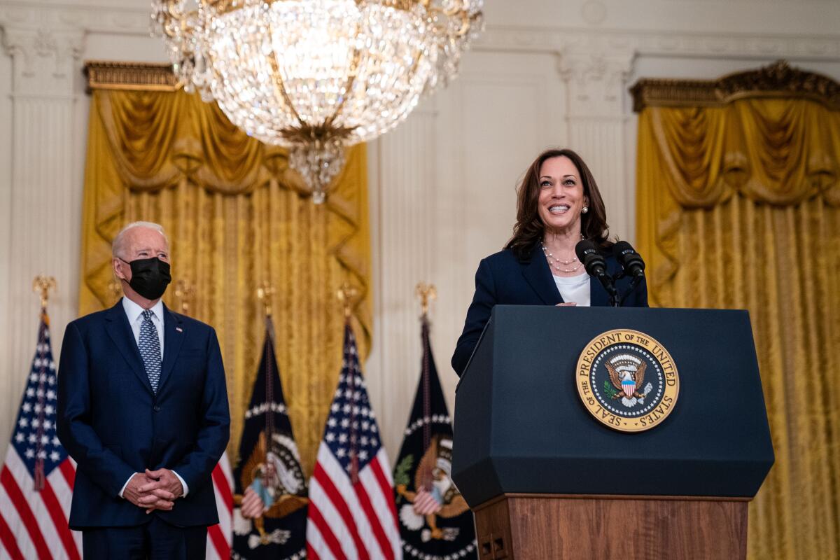 Kamala Harris speaks at a White House lectern while Joe Biden, in a face mask, stands behind and to the side.