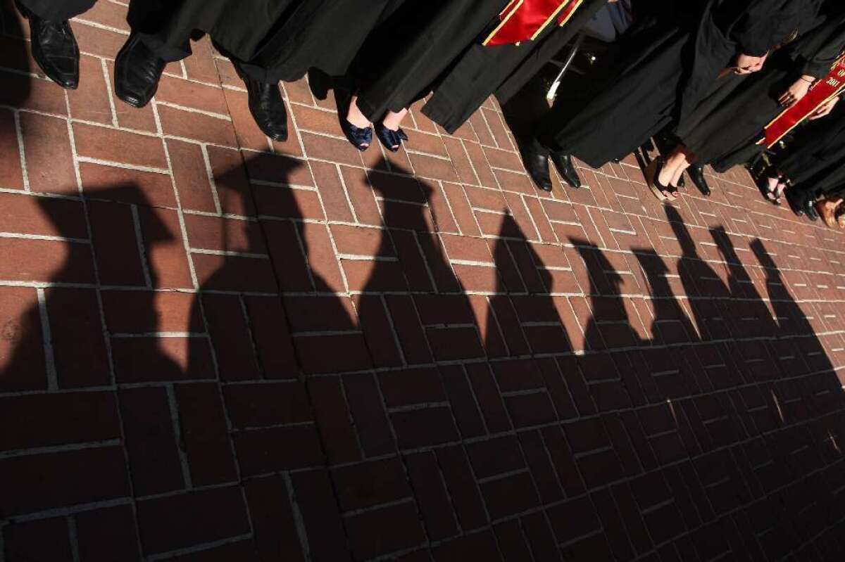 Shadows of students in caps and gowns on paving bricks near other people in black graduation gowns.