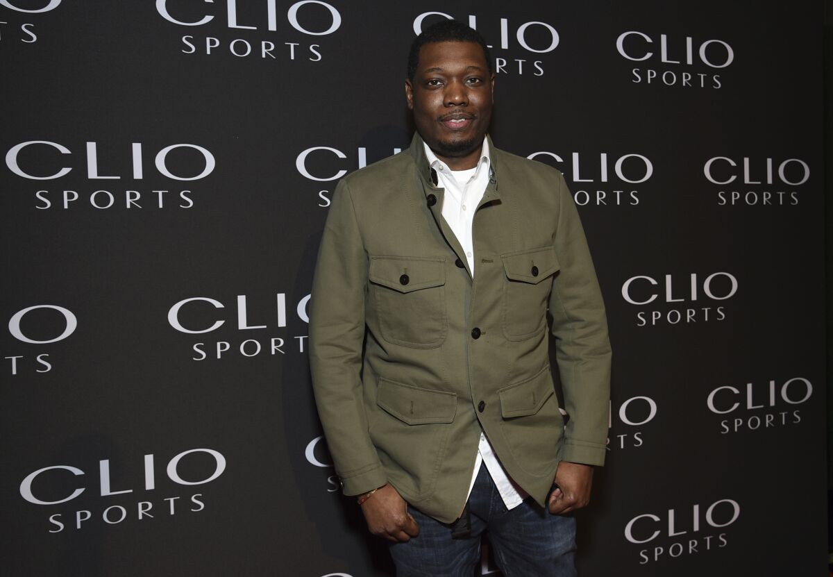 A man posing in a green jacket, white shirt and jeans against a black backdrop