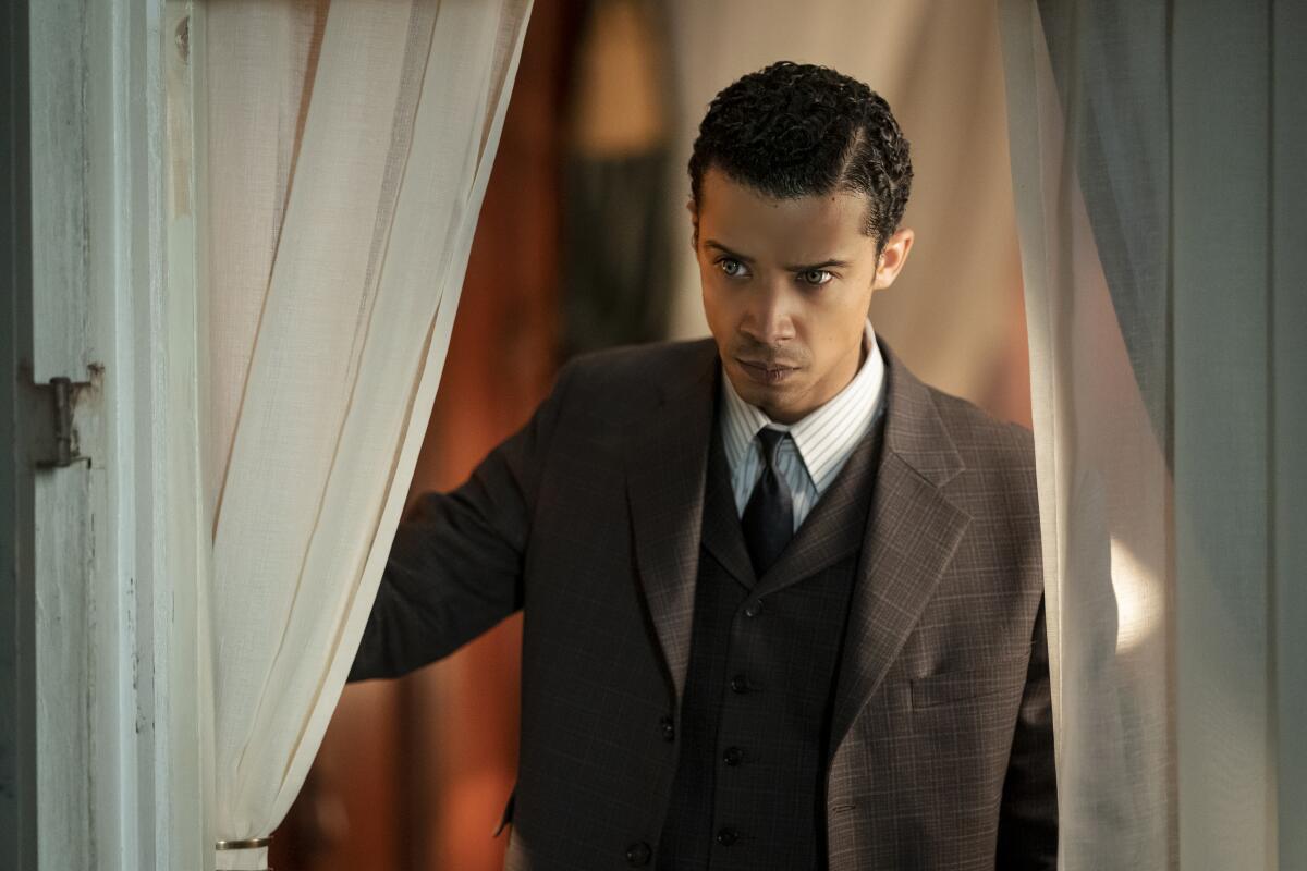 A man in a dapper suit holds back the curtains while looking through the window.