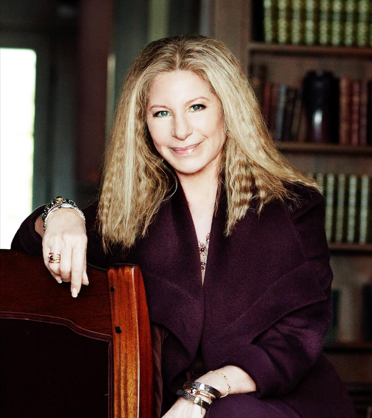 Barbra Streisand rests her arm on the back of the wooden chair she is sitting in for a portrait.