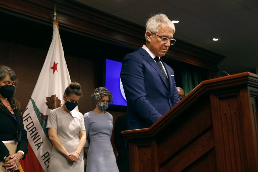 Los Angeles, California-Dec. 8, 2021-Los Angeles District Attorney George Gascon, right, bows his head at the beginning a press conference after asking others to observe a moment of silence in honor of the victims of recent shootings in Los Angeles. Supporting him during his press conference were from left to right are Miriam Krinsky, left, Laura Conover, second from left, and Parisa Tafti, third from left. (Carolyn Cole / Los Angeles Times)