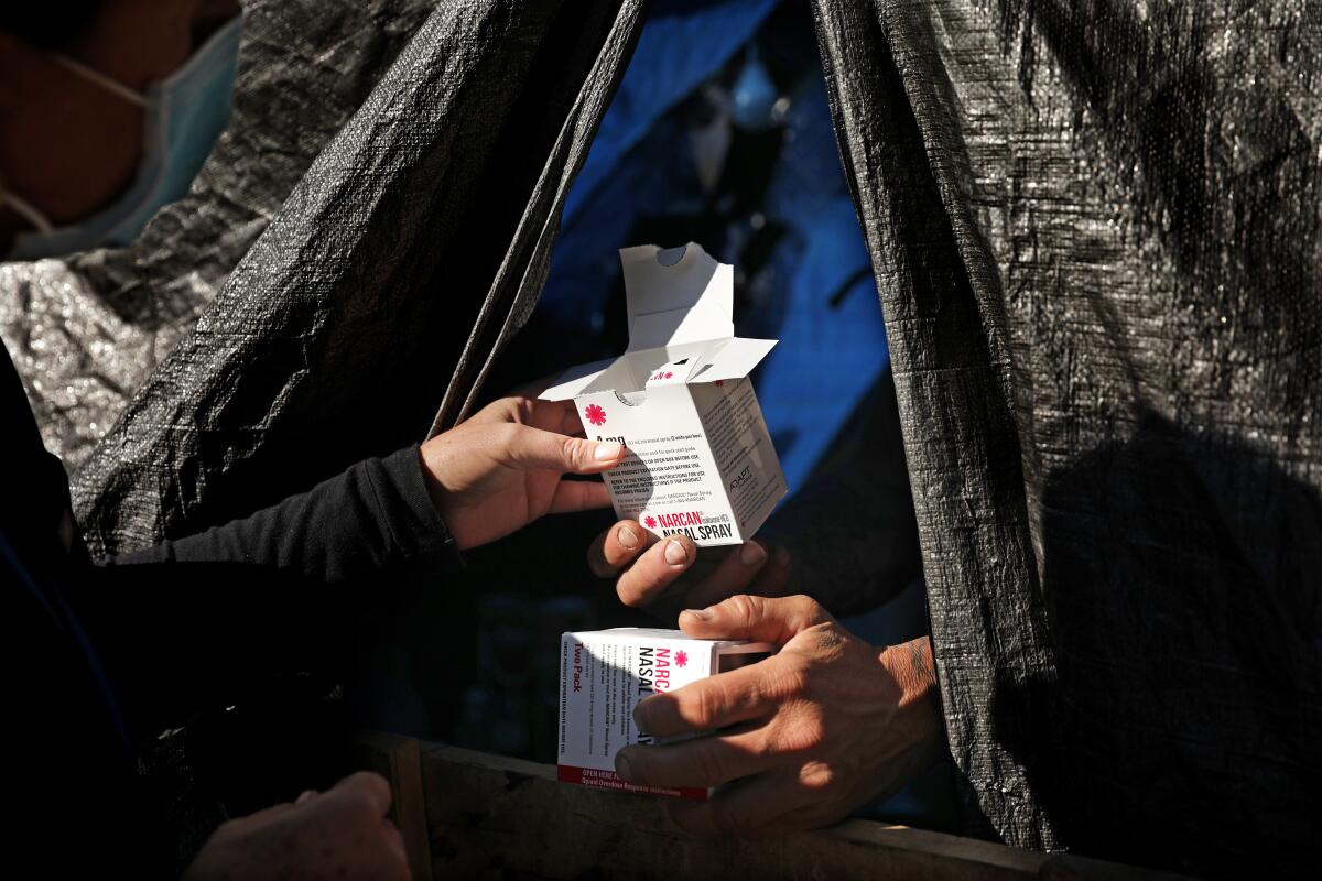 A hand offers a box of Narcan nasal spray to a man living in a makeshift structure