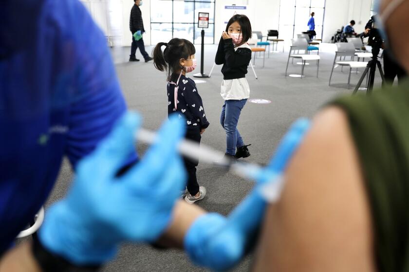 SANTA ANA-CA-APRIL 22, 2021: Chloe Choi, 7, right, takes a peek as her sister Bella, 4, left, turns away as they try not to watch their dad receive his first shot at a new mass vaccination site in Orange County-the Providence Vaccine Clinic at Edwards Lifescience in Santa Ana on Thursday, April 22, 2021. (Christina House / Los Angeles Times)
