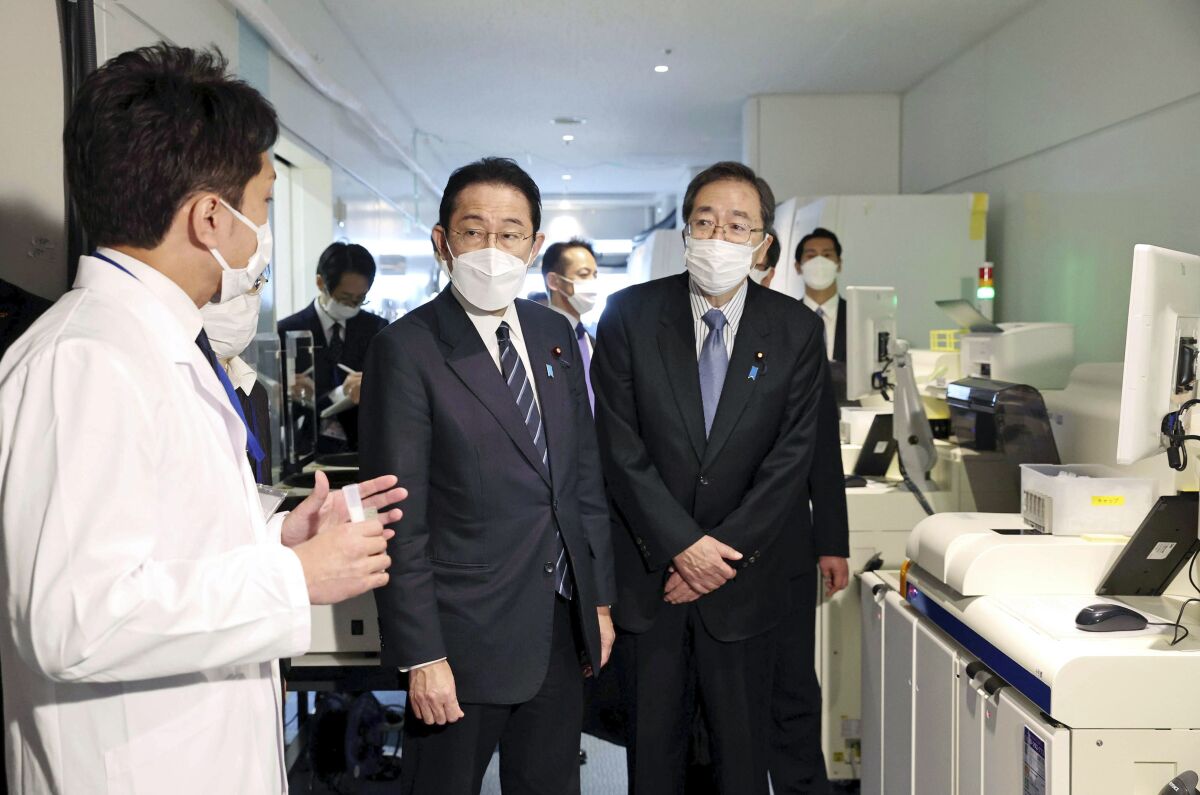 Japanese Prime Minister Fumio Kishida, center, listens to an official at an quarantine area at the Haneda international airport in Tokyo, Saturday, Feb. 12, 2022. Japan is considering easing its stringent border controls amid growing criticism that the measures, which have banned most foreign entrants including students and business travelers, are hurting the country's economy and international profile. (Japan Pool/Kyodo News via AP)