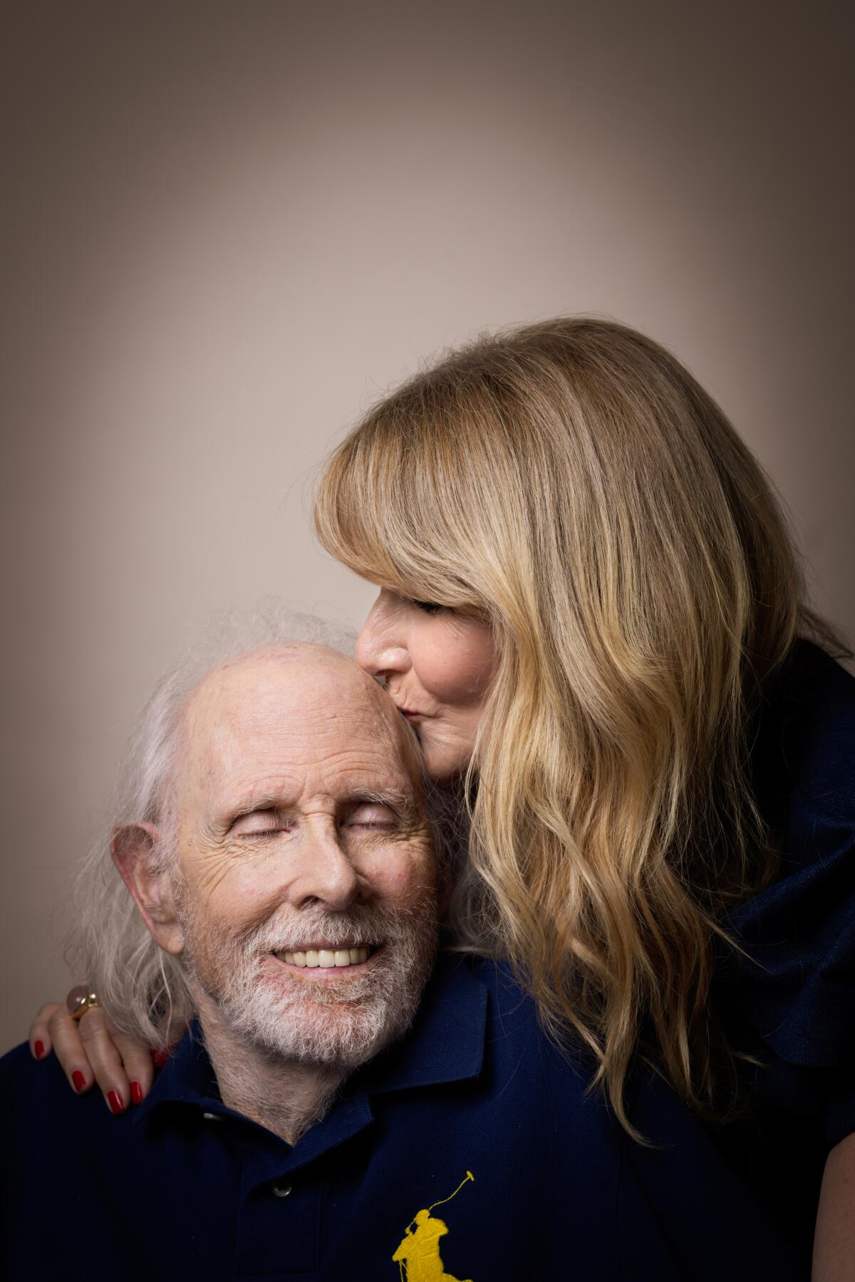 A blond woman kisses the top of the head of an older man with white hair and a white beard.