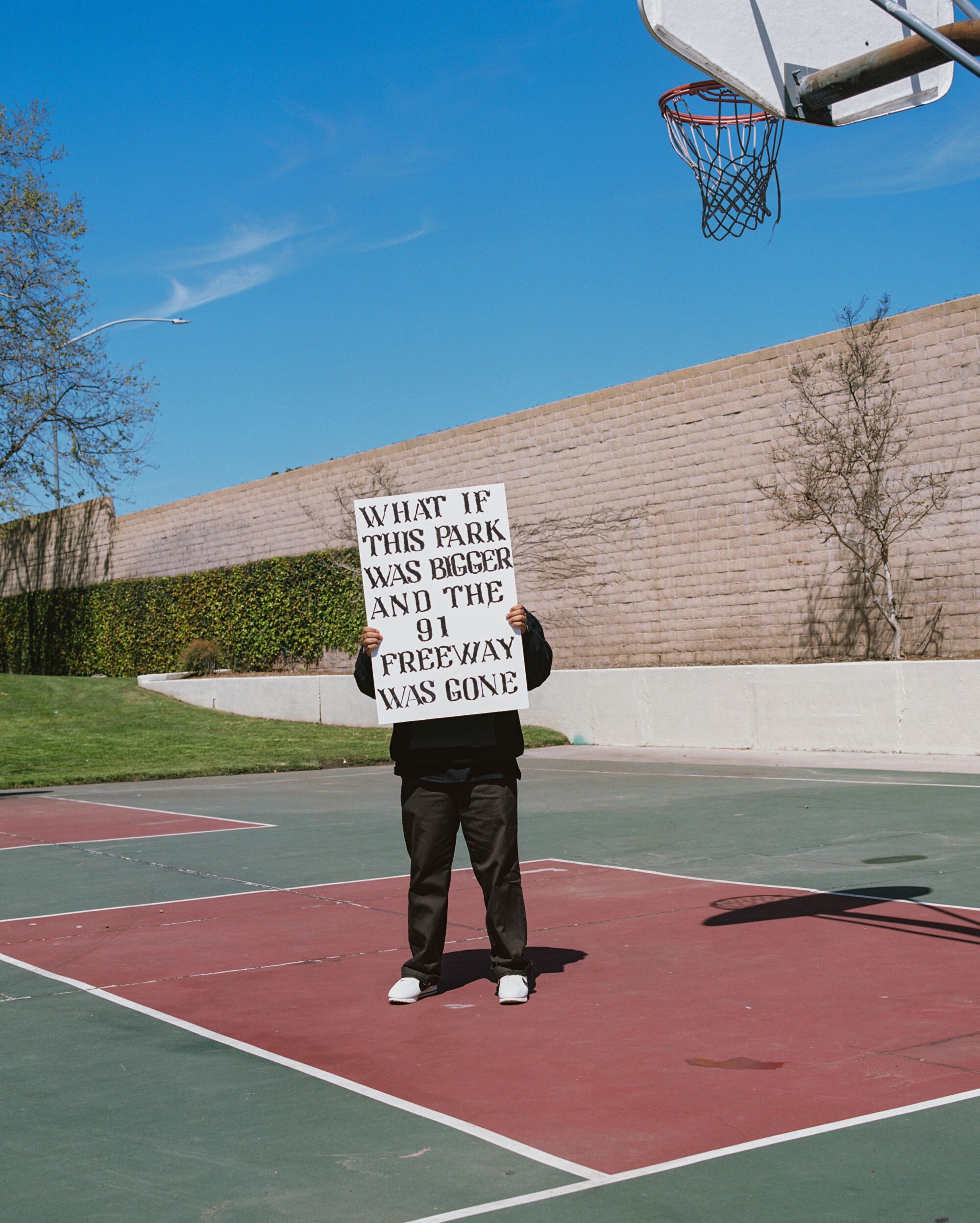 A man stands on a basketball court with the sign: "What If This Park Was Bigger and the 91 freeway as gone?"