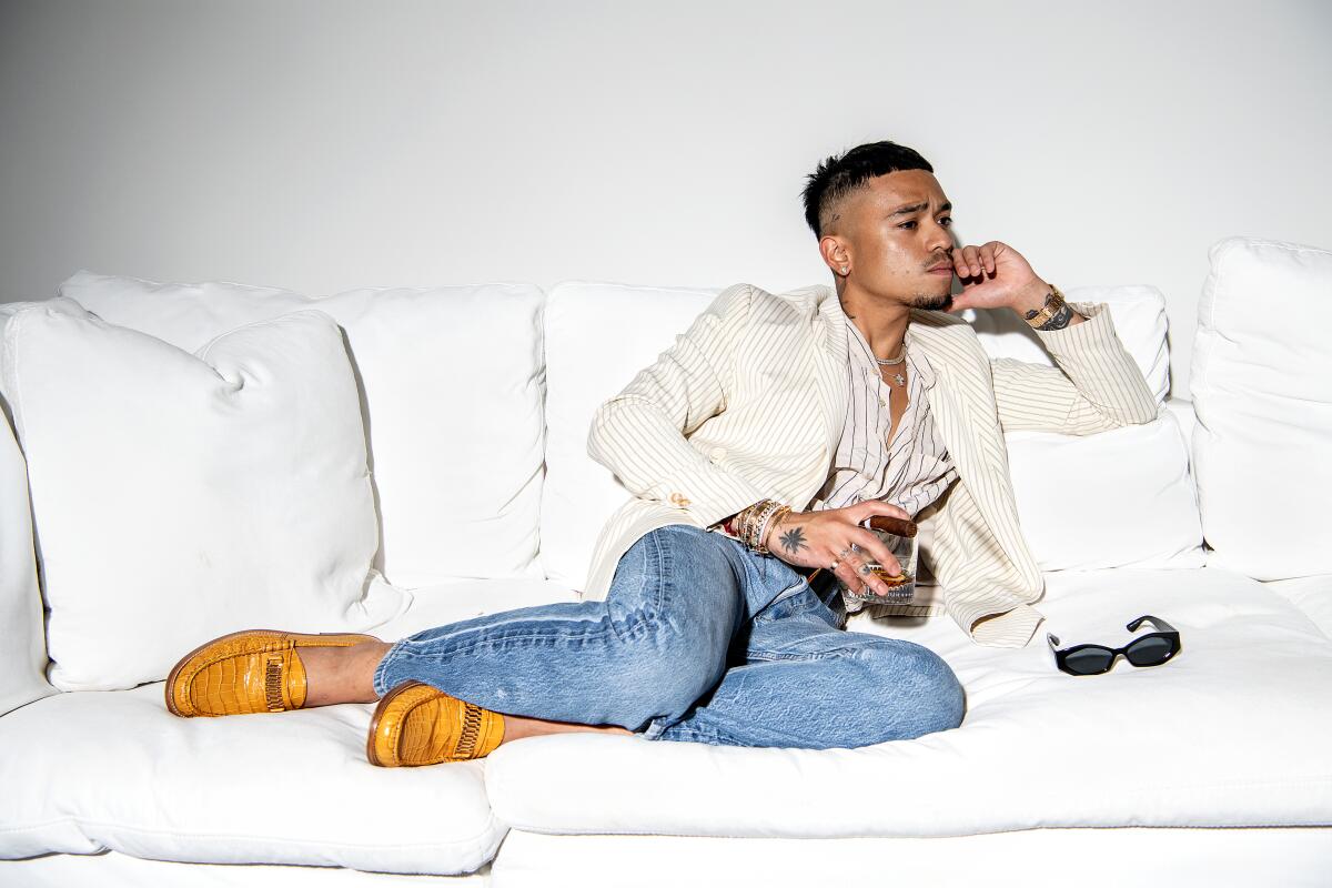 Rhuigi Villasenor, in jeans, loafers and white suit coat, lounges on a white couch with drink in hand.