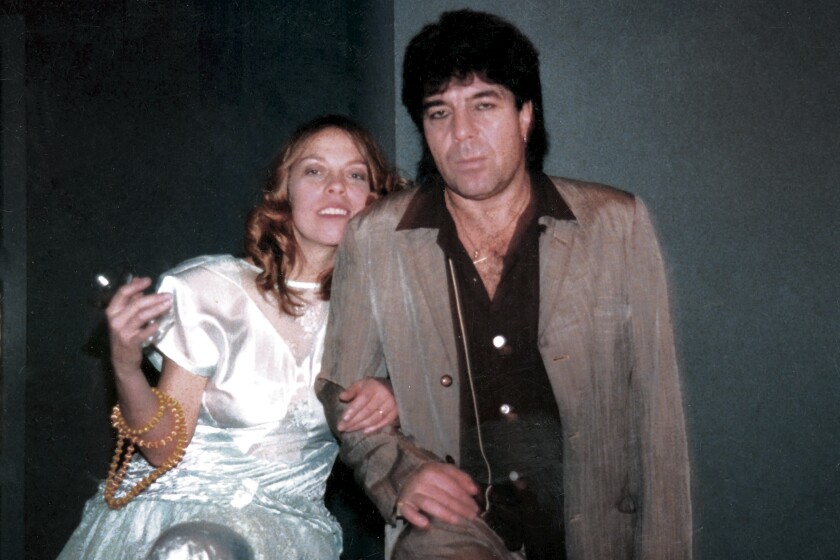 A woman in a white silk dress, left, leans against a man wearing a brown coat and brown shirt