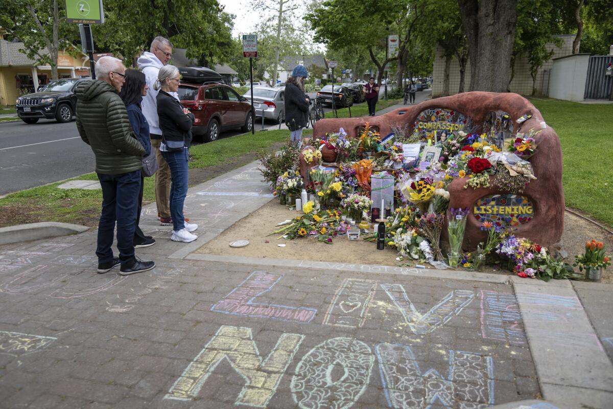 People stand on a sidewalk looking at flowers and other tributes on a free-form bench that reads "Compassion" and "Listening"