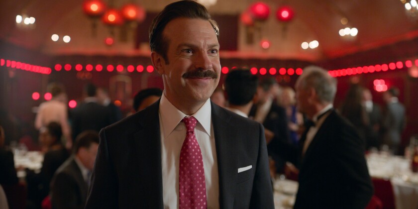 Jason Sudeikis as "Ted Lasso," in a navy suit with red tie.