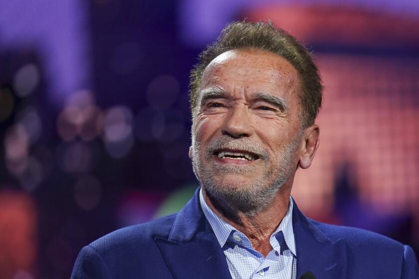 Arnold Schwarzenegger with gray facial hair smiling in a blue suit shirt and blazer