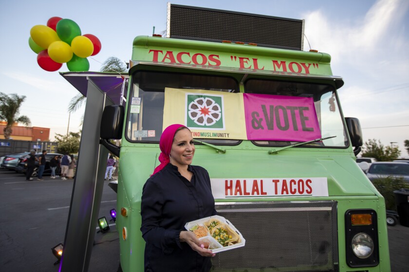 A smiling woman with a plate of tacos in front of a truck with banners including "halal tacos."