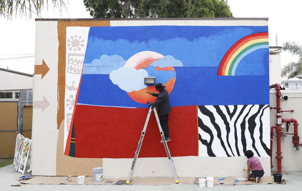 Michael Massenburg stands on a ladder, making repairs to a bright red and blue mural that features a rainbow.