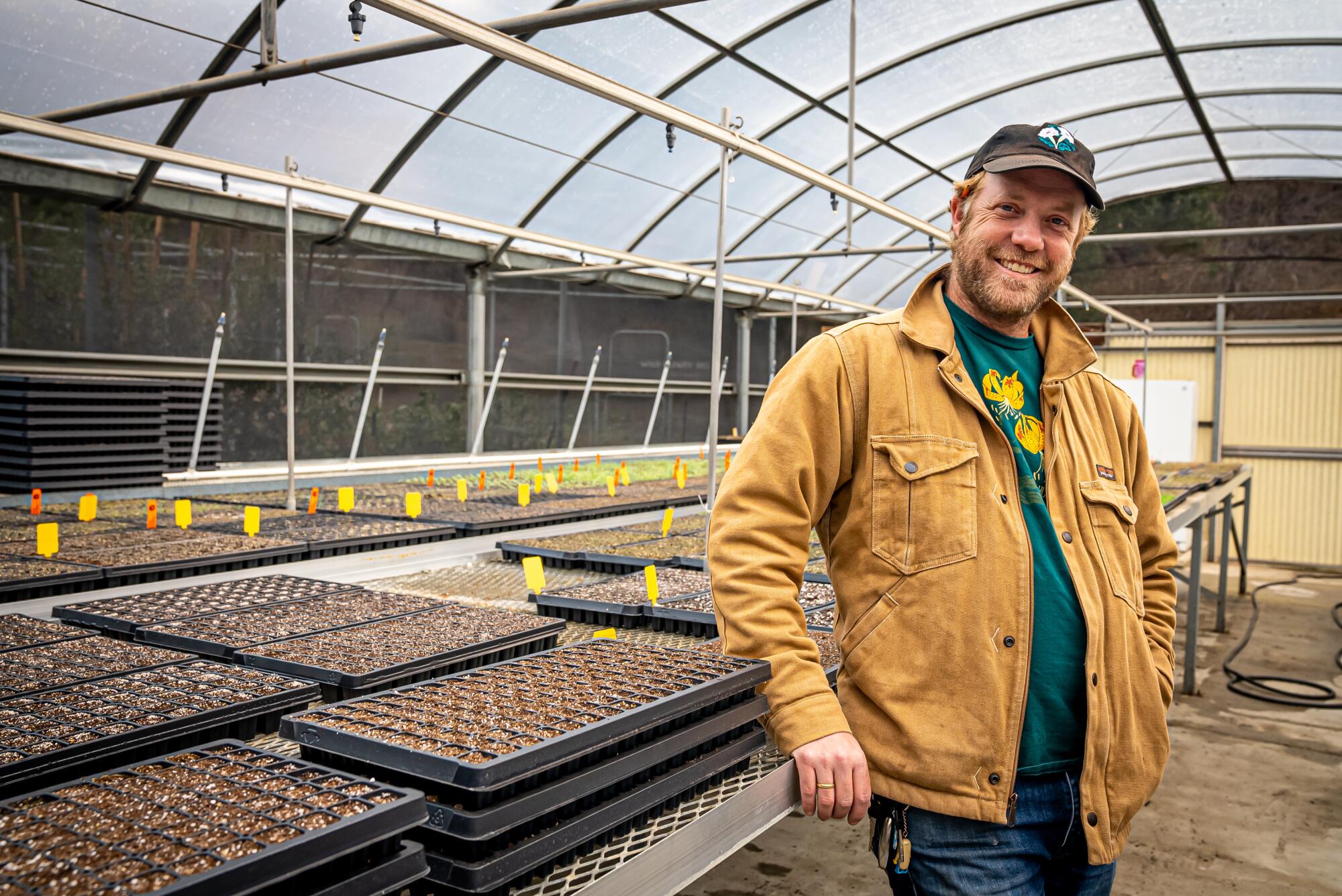 A smiling man stands next to trays of seedlings in a greenhouse.