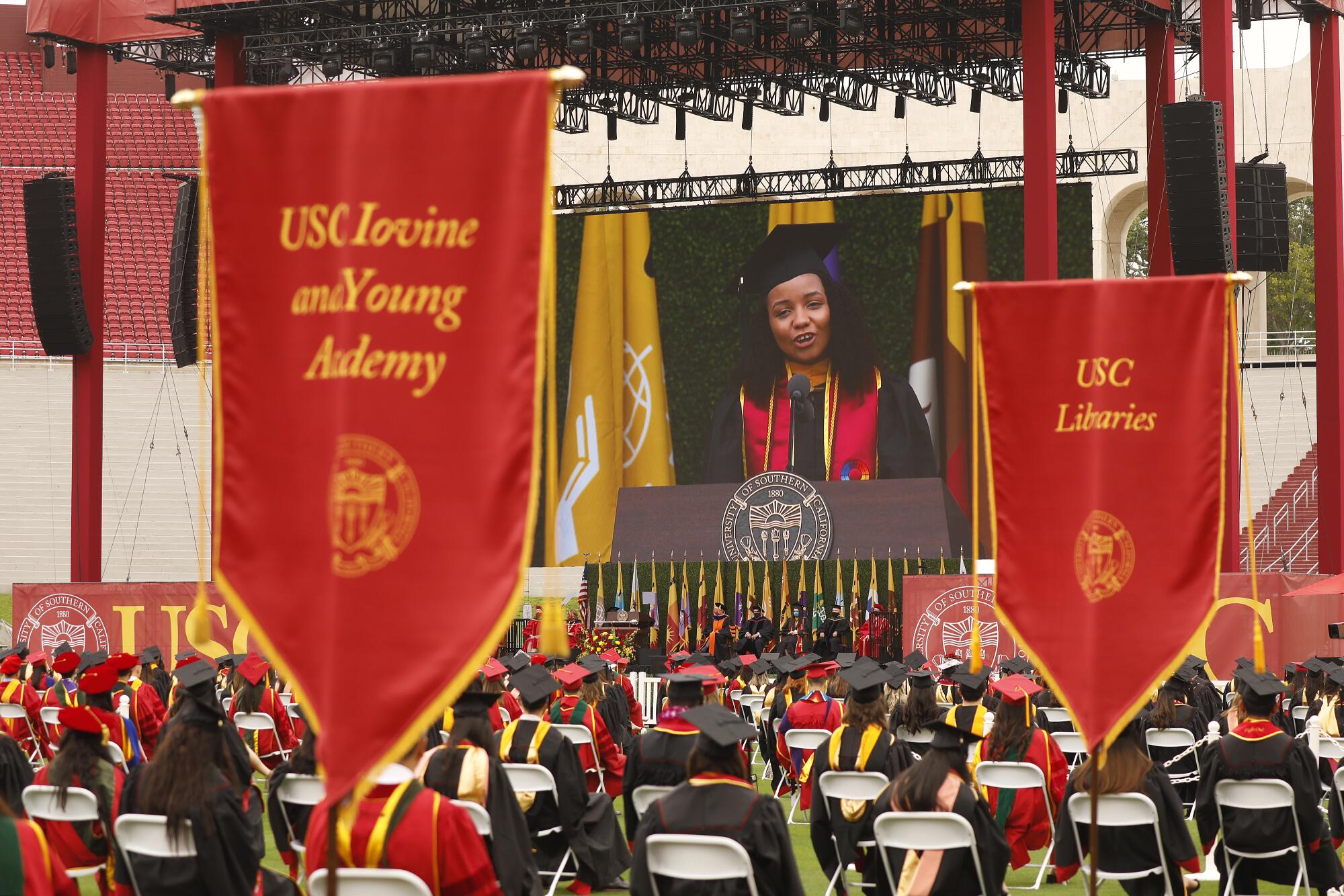 A large screen behind the graduation stage shows a student in a black gown and red sash giving a speech