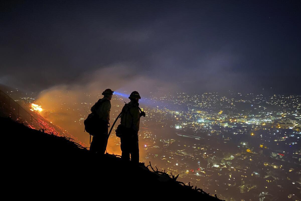 Two people in silhouette on a mountain appear to look at a fire on the left and a city on the right full of lights at night.
