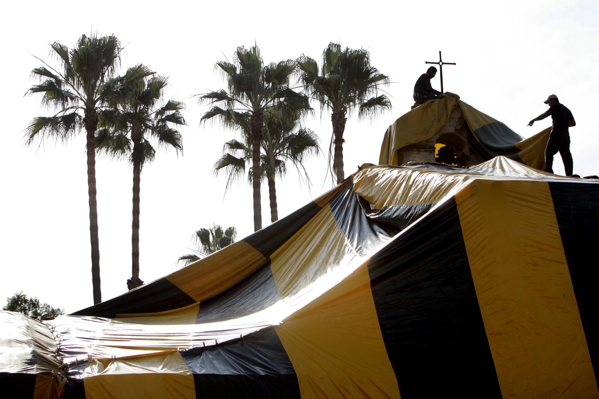 Workers drape a black and yellow fumigation tent over a building.