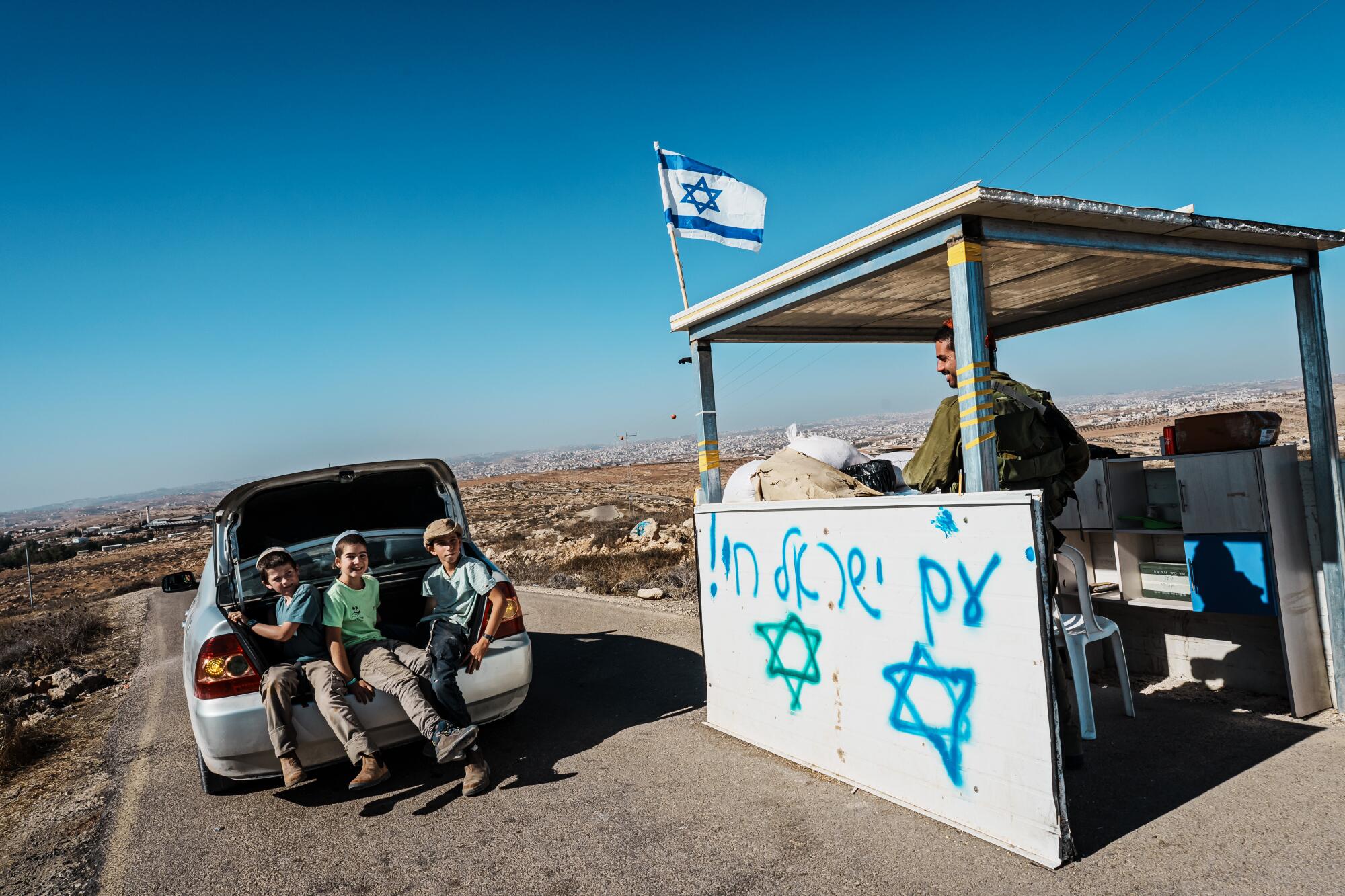 A man in a roadside stand with a blue-and-white flag looks at three young people sitting in the open trunk of a car 