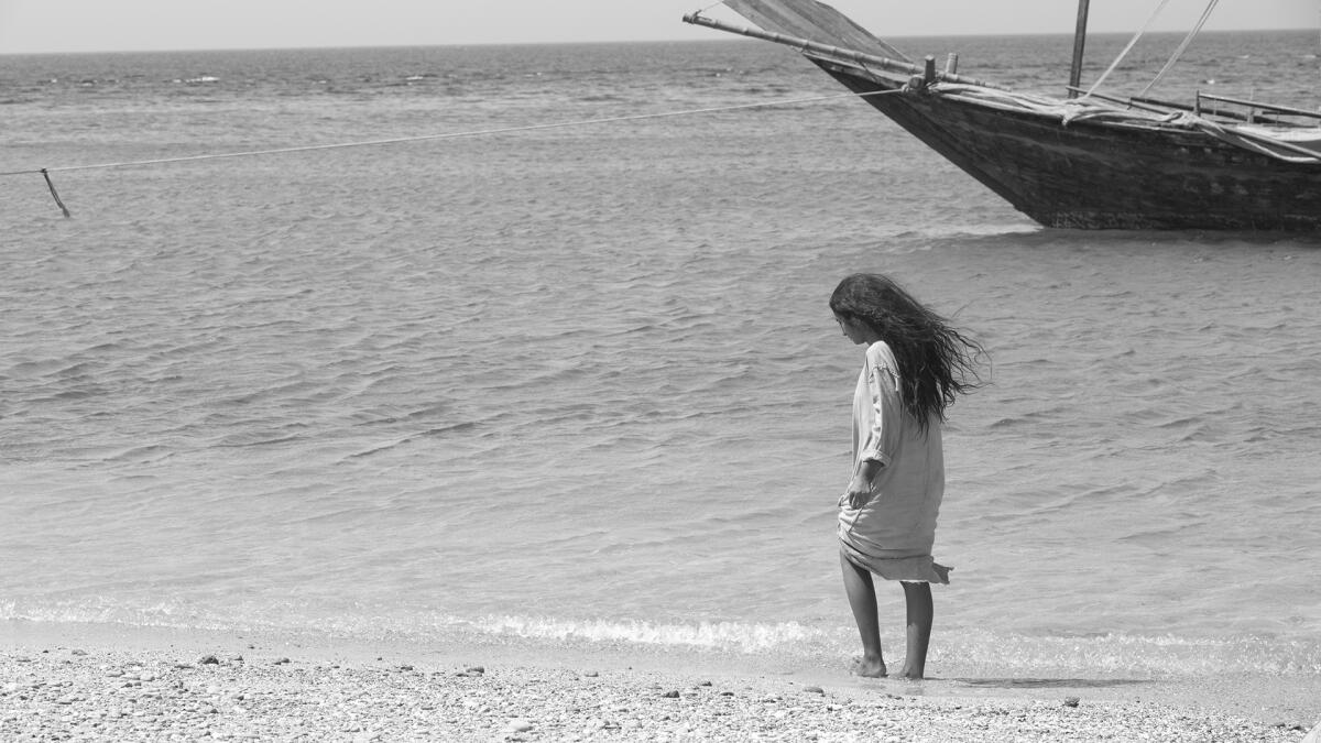 A girl walks along a shoreline with a boat in the background in the movie "Scales."