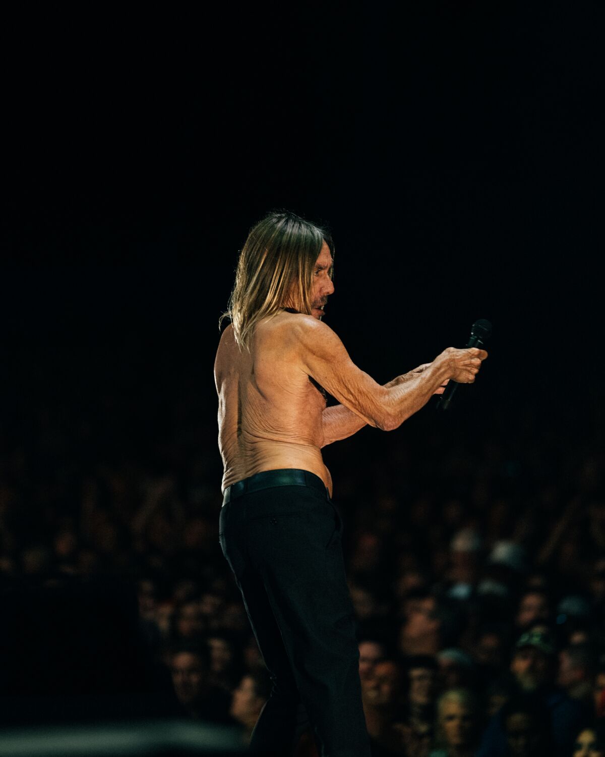 A shirtless man with long hair performs onstage before a crowd.