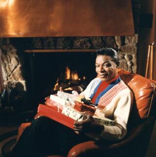 Nat King Cole sits by a fireplace holding a stack of gifts.
