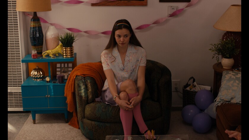A young woman sits on a green swivel chair with pink streamers in the background.