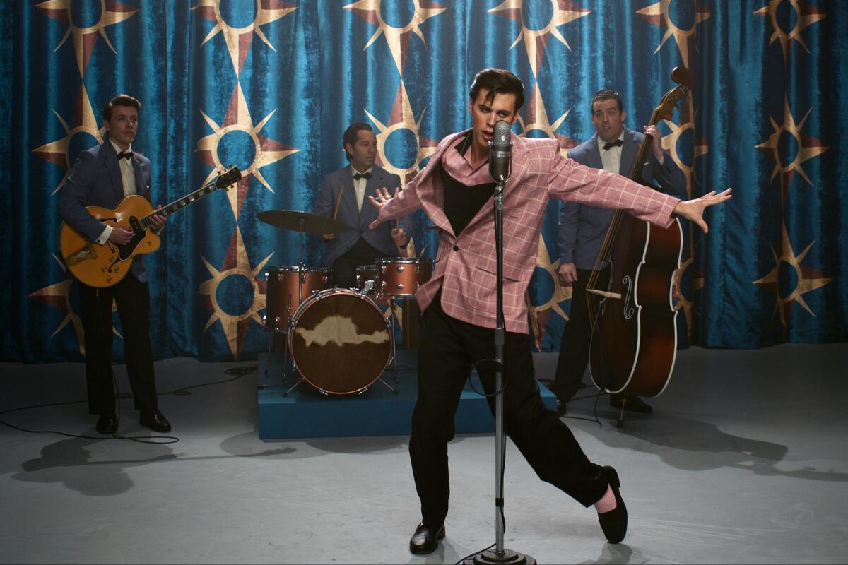 A man in a pink jacket at a microphone strikes a pose before a band in the movie "Elvis."