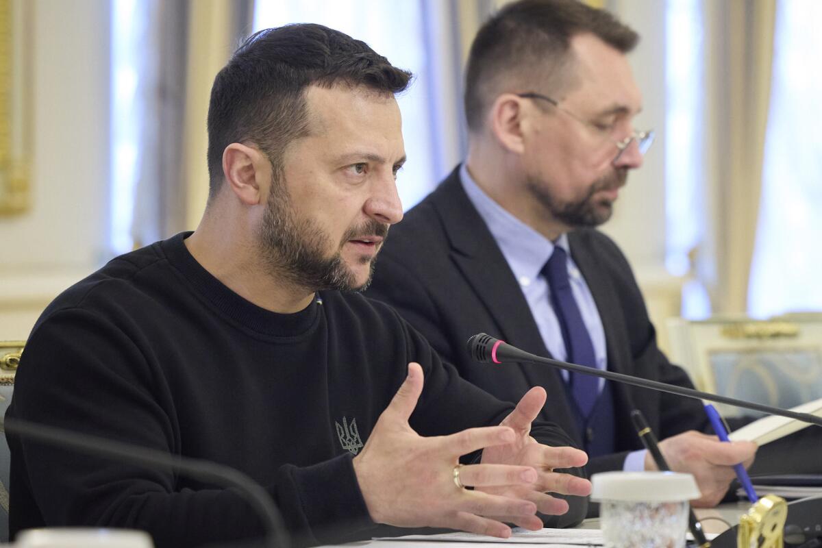 A man in a black sweater sits at a table and speaks into a small microphone as he gestures with his hands.