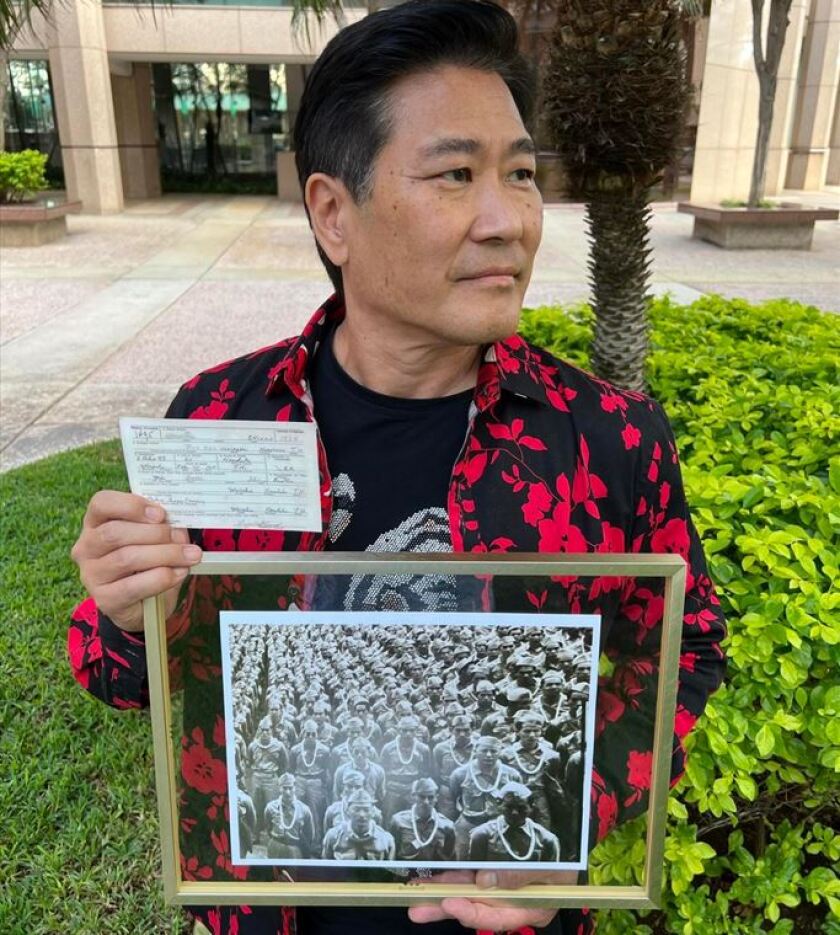 Rob Shinno holds photos of his dad’s draft card and his dad amid a large group of troops in Honolulu.