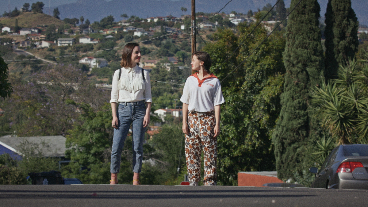 Two women stand in a Los Angeles street with hillside homes behind them.