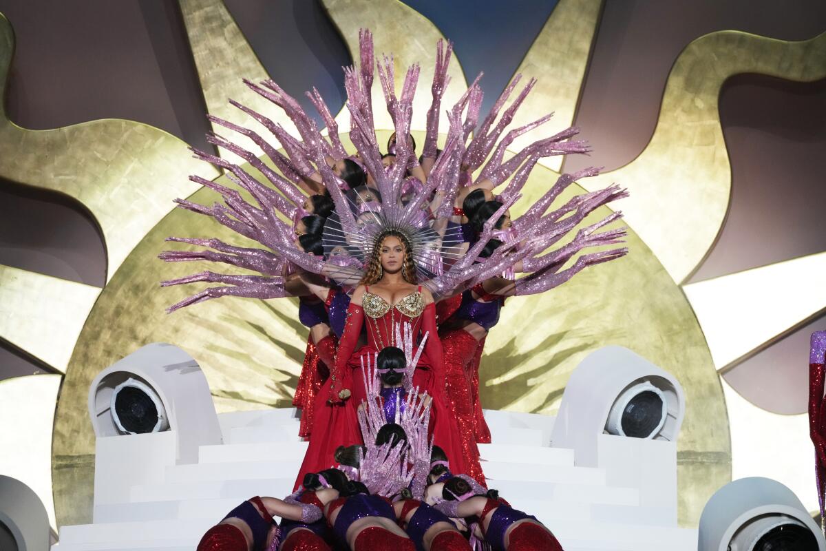 Beyoncé, in a glittering gown and surrounded by dancers, stands in front of a large sun design