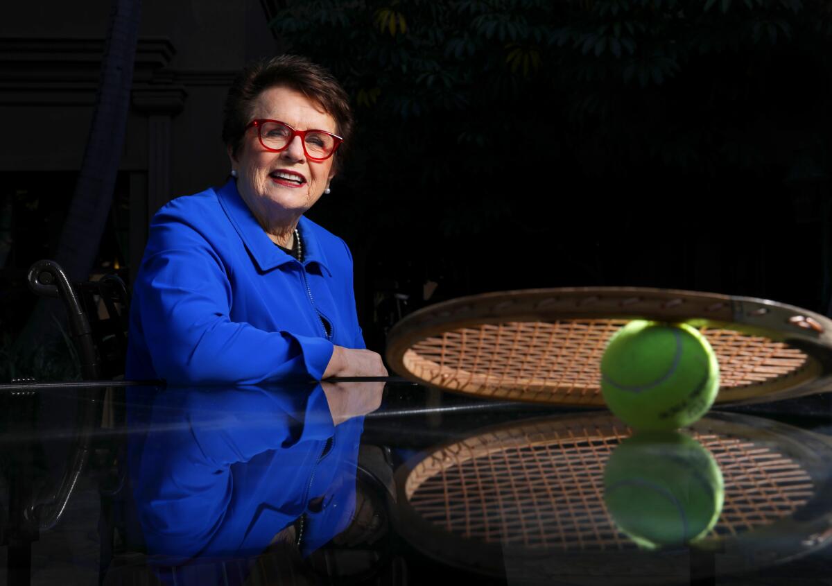 Billie Jean King sits at a table with a tennis racket.