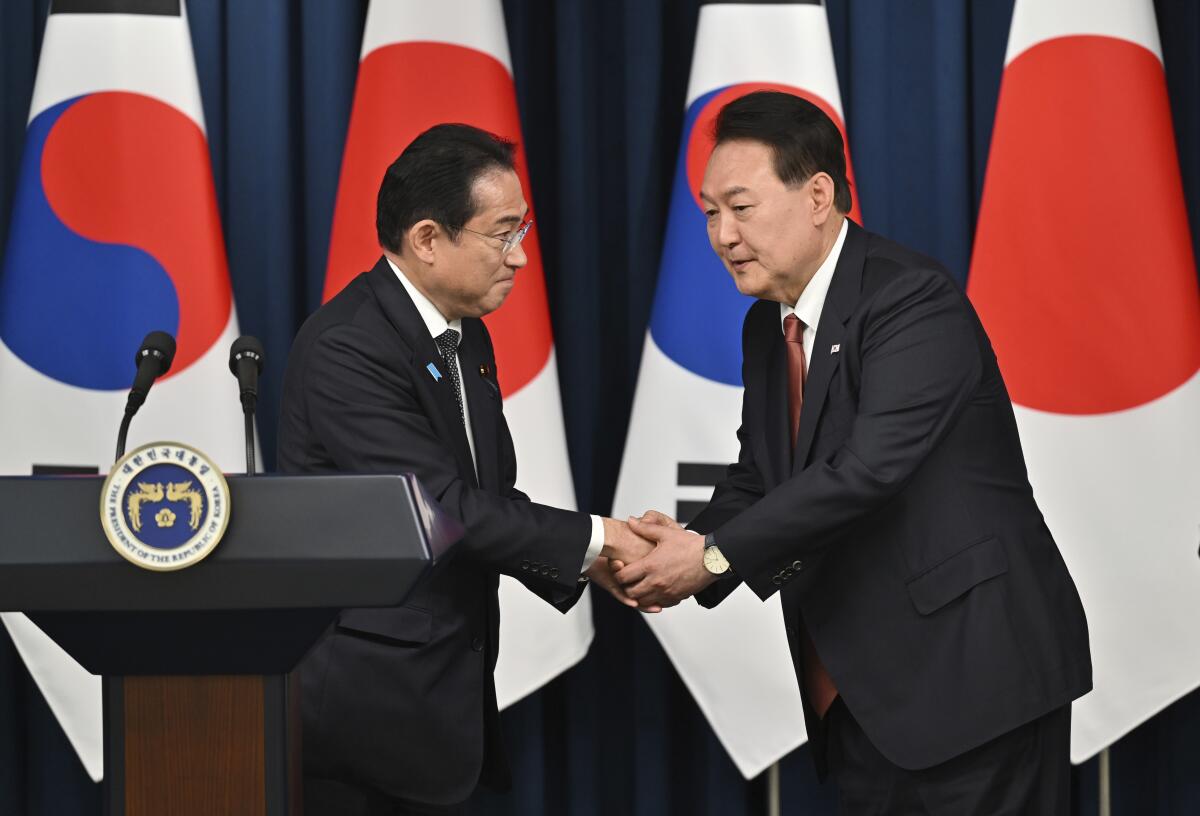 South Korean President Yoon Suk Yeol shakes hands with Japanese Prime Minister Fumio Kishida in front of flags.