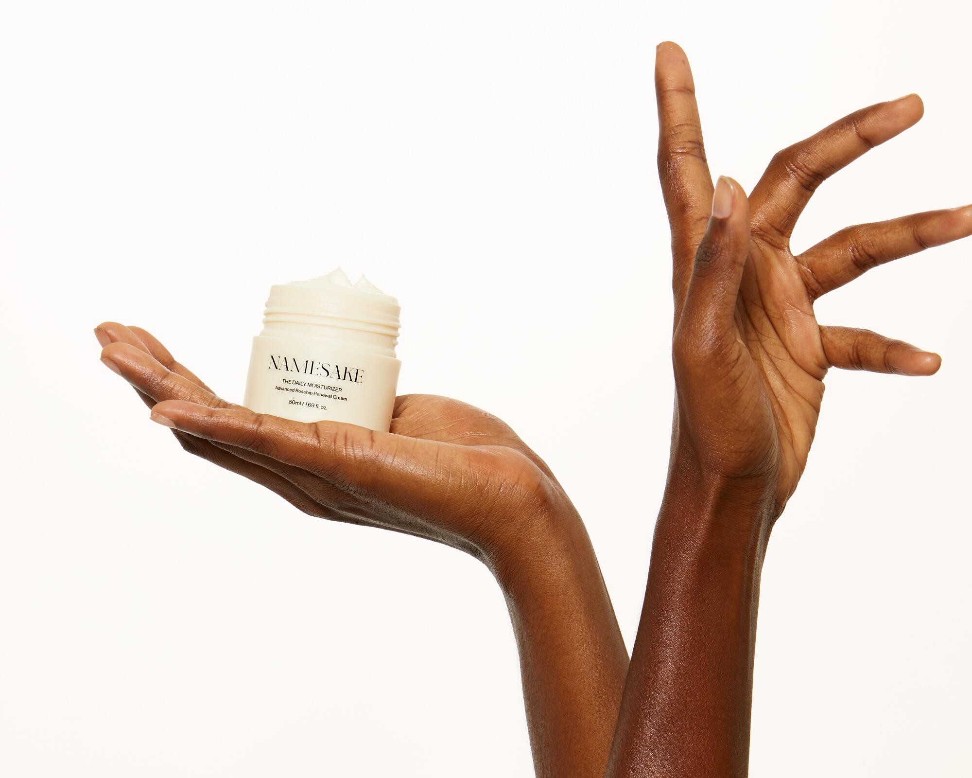 a pair of brown hands in the air on a white background, with one hand cupping a jar of Namesake daily moisturizer
