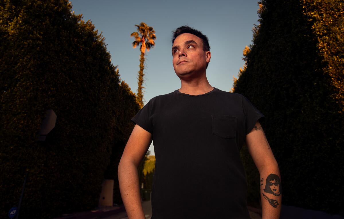 A man with a tattoo on his arm and wearing a black T-shirt stands in front of hedges and a palm tree.