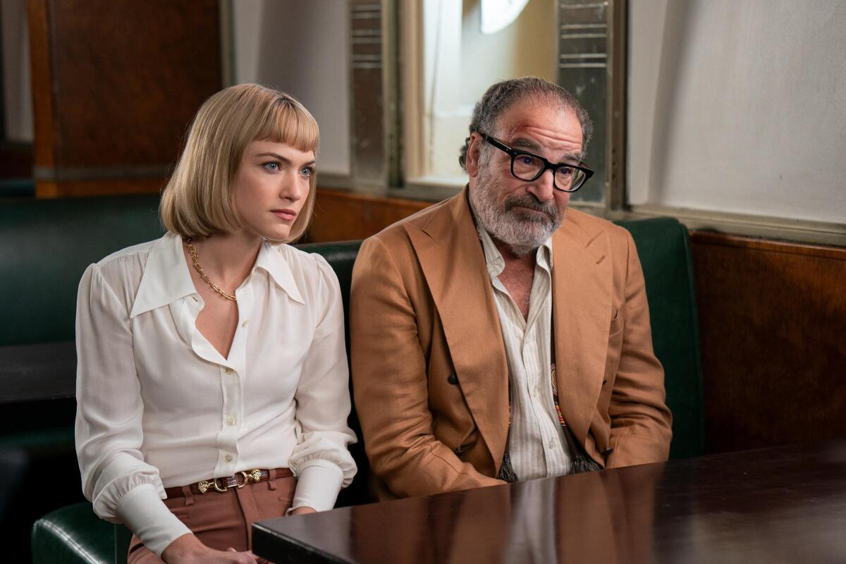 A blonde woman in a white shirt and an older man in glasses, a brown blazer and white shirt sit next to each other.