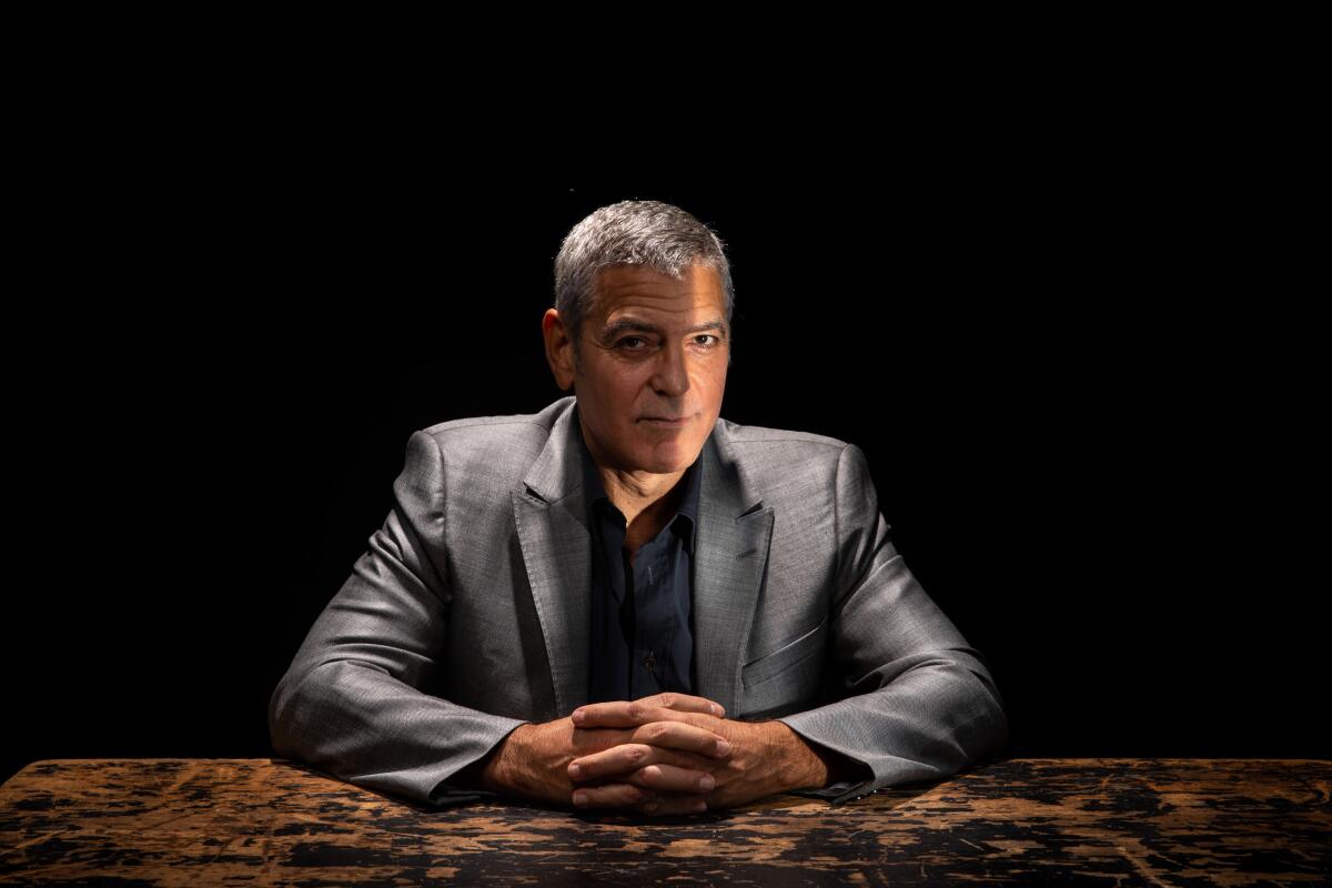 George Clooney sits at a table in a gray suit with hands clasped